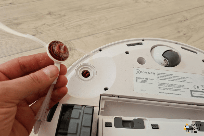 Die Seitenbürste des ECOVACS DEEBOT T10 wird abgenommen und die sich dort aufgewickelten Haare werden mit der Hand entfernt.